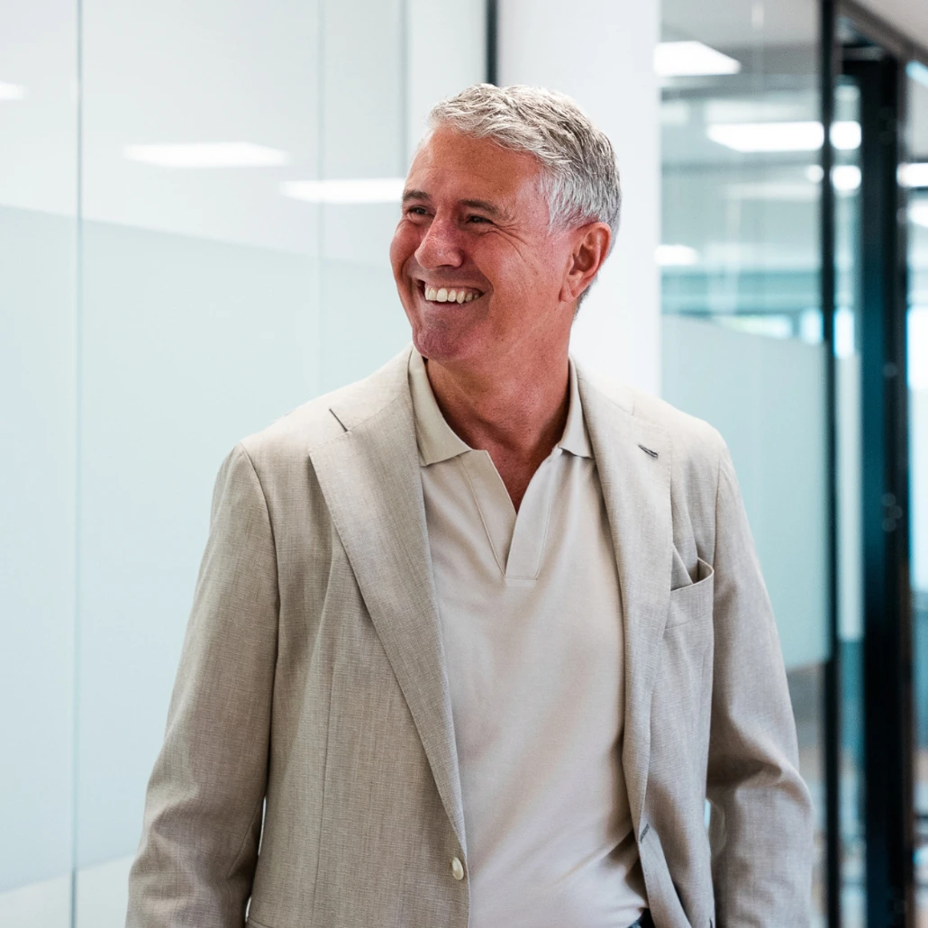 Enrico Ouwenhand lacht en kijkt opzij in een modern kantoorgebouw bij Salesrecruiters, gekleed in een lichtgrijs jasje met een poloshirt eronder.